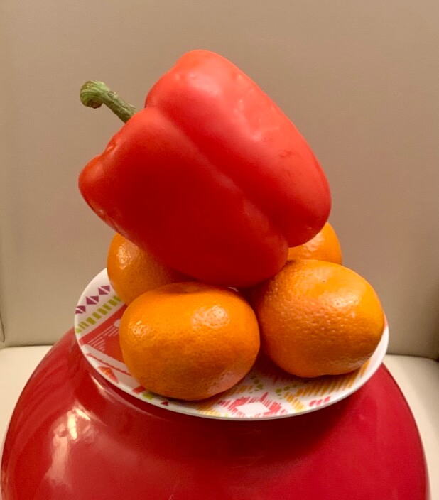 A red pepper balances on a pile of oranges sitting on a plate